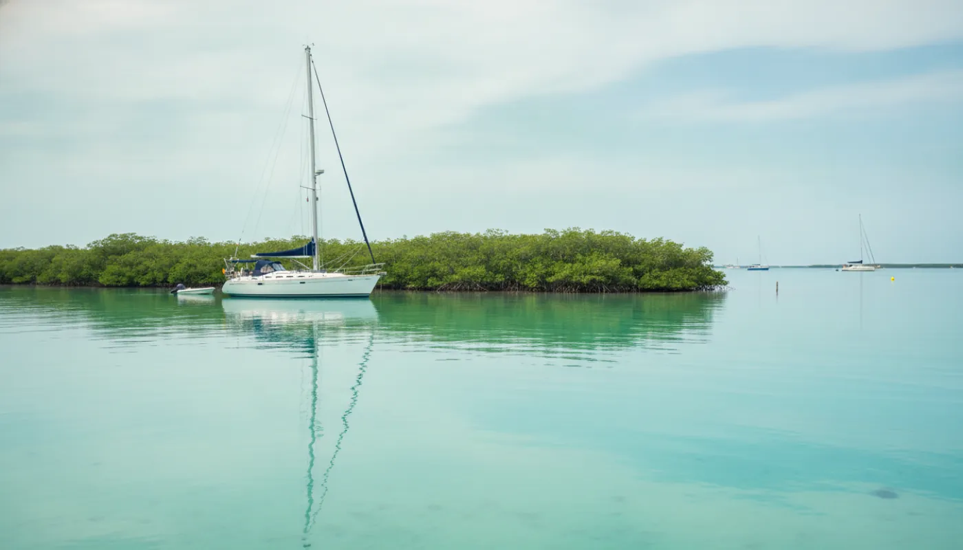 A cruising sailboat anchored in shallow turquoise water off a mangrove-fringed Florida Keys island at midday, white hull mirrored on the calm surface, low green keys and a few distant boats under a bright sky